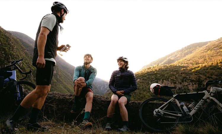 grupo de três ciclistas conversando e sorrindo com as bicicletas estacionadas com montanhas ao fundo